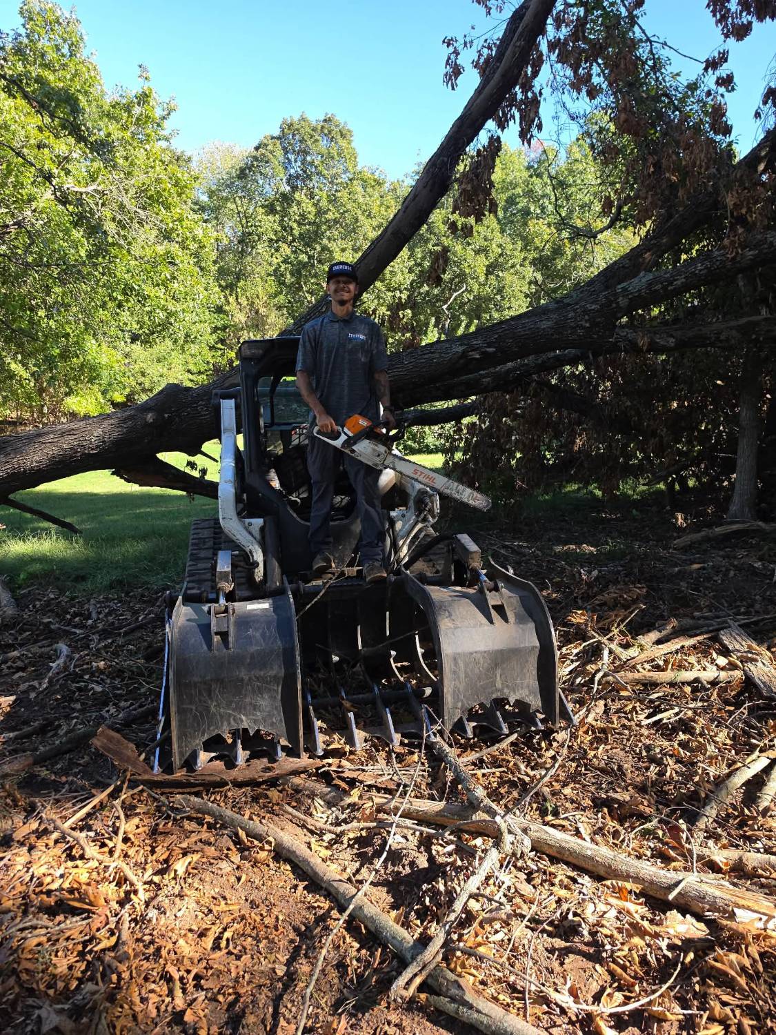 Byron, owner of Ever Edge Outdoor Solutions, holding a chainsaw on a track loader in front of a fallen tree